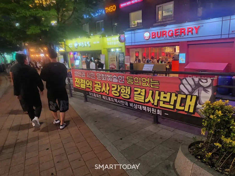 행인들이 대장홍대선 레드로드 역사 설치 반대 현수막 옆 거리를 지나고 있다. 출처=김종현 기자