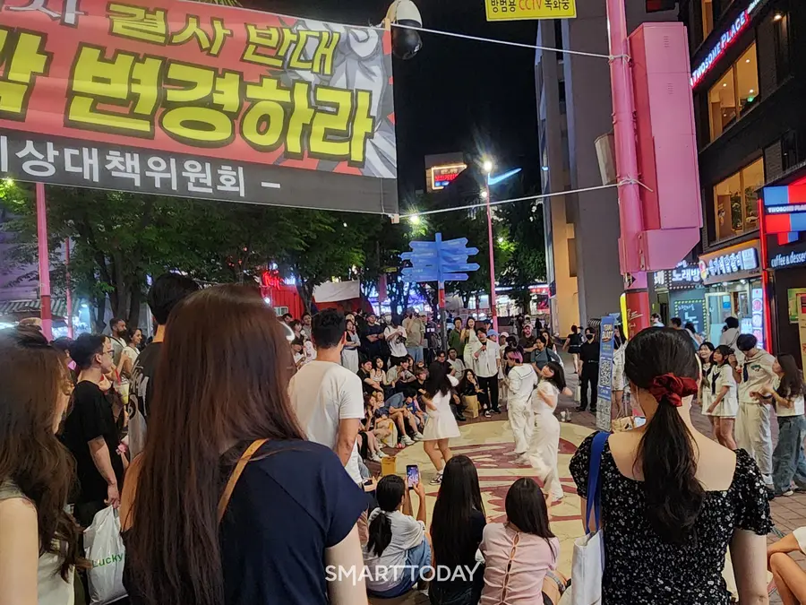 관광객들이 버스킹 춤 공연을 관람하고 있다. 대장홍대선 역사 건설 반대 현수막이 인근에 설치돼 있다. 출처=김종현 기자